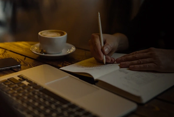 Person writing in notebook at café table with coffee and laptop