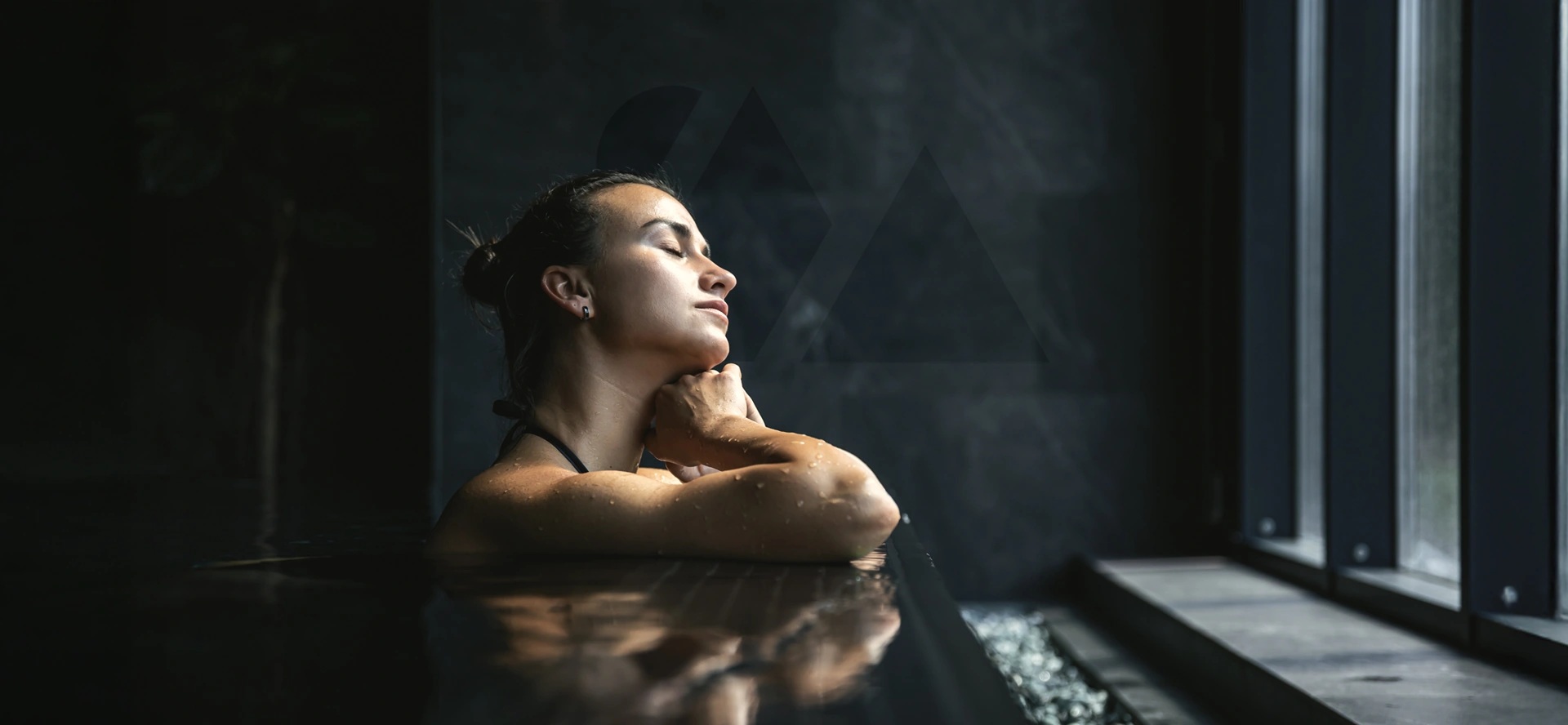 Woman relaxing in indoor hot spring pool with eyes closed and natural light